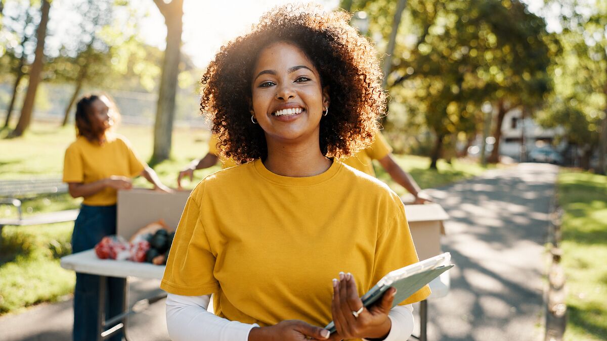 A young volunteer in a yellow programme T-shirt smiling to camera, clipboard in hand, at an outdoor community drive